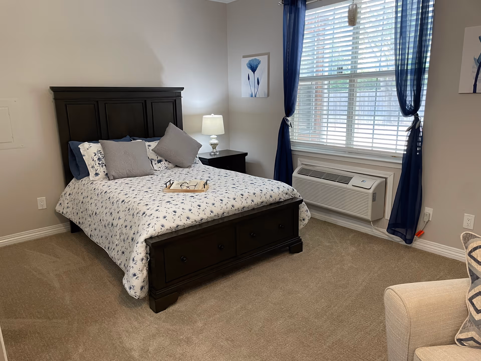 A bedroom with a dark wooden bed frame and a floral-patterned bedspread in white and blue. There are two gray pillows and two blue pillows on the bed, along with a tray holding a pair of glasses and a book. A nightstand with a lamp is next to the bed. The room has beige carpet, a window with blue curtains, and an air conditioning unit below the window. A beige armchair with a patterned pillow is partially visible in the foreground.