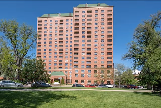 A tall brick residential building with multiple windows and green rooftops, surrounded by trees and parked cars on a sunny day with a clear blue sky.