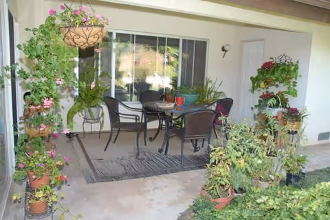 Covered outdoor patio area with a table and four chairs on a rug, surrounded by various potted plants and hanging flower baskets, adjacent to a building with a window and a door.
