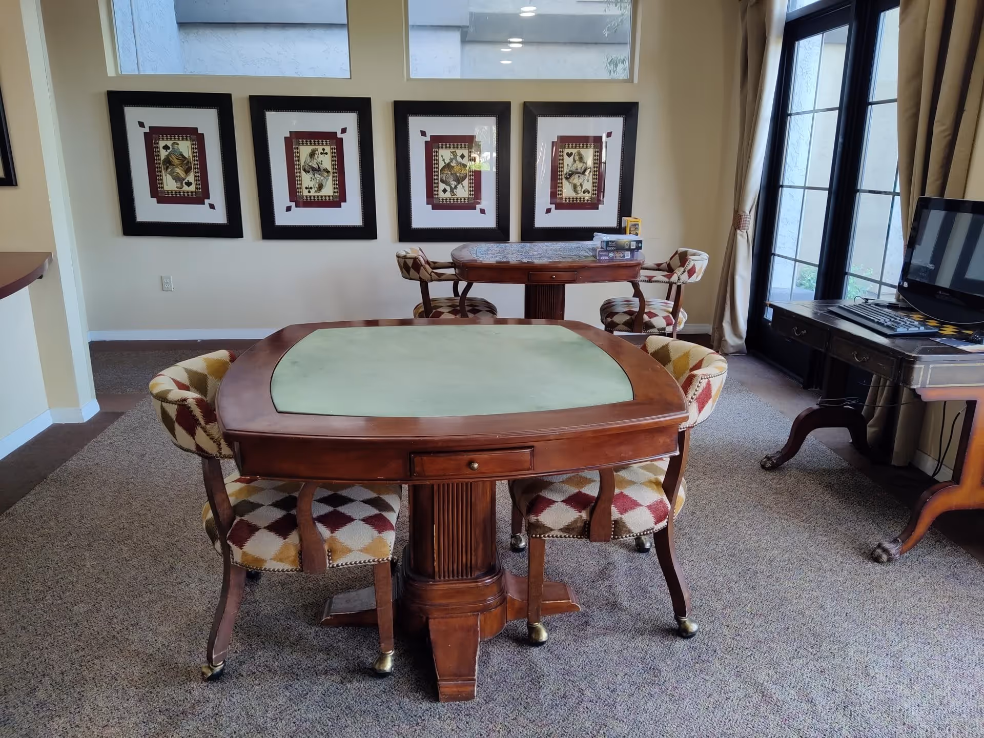 A cozy interior room with two wooden tables, each surrounded by four cushioned chairs with a diamond pattern upholstery. The room has carpeted flooring, framed artwork on the wall, and a computer desk with a monitor near a glass door with curtains.