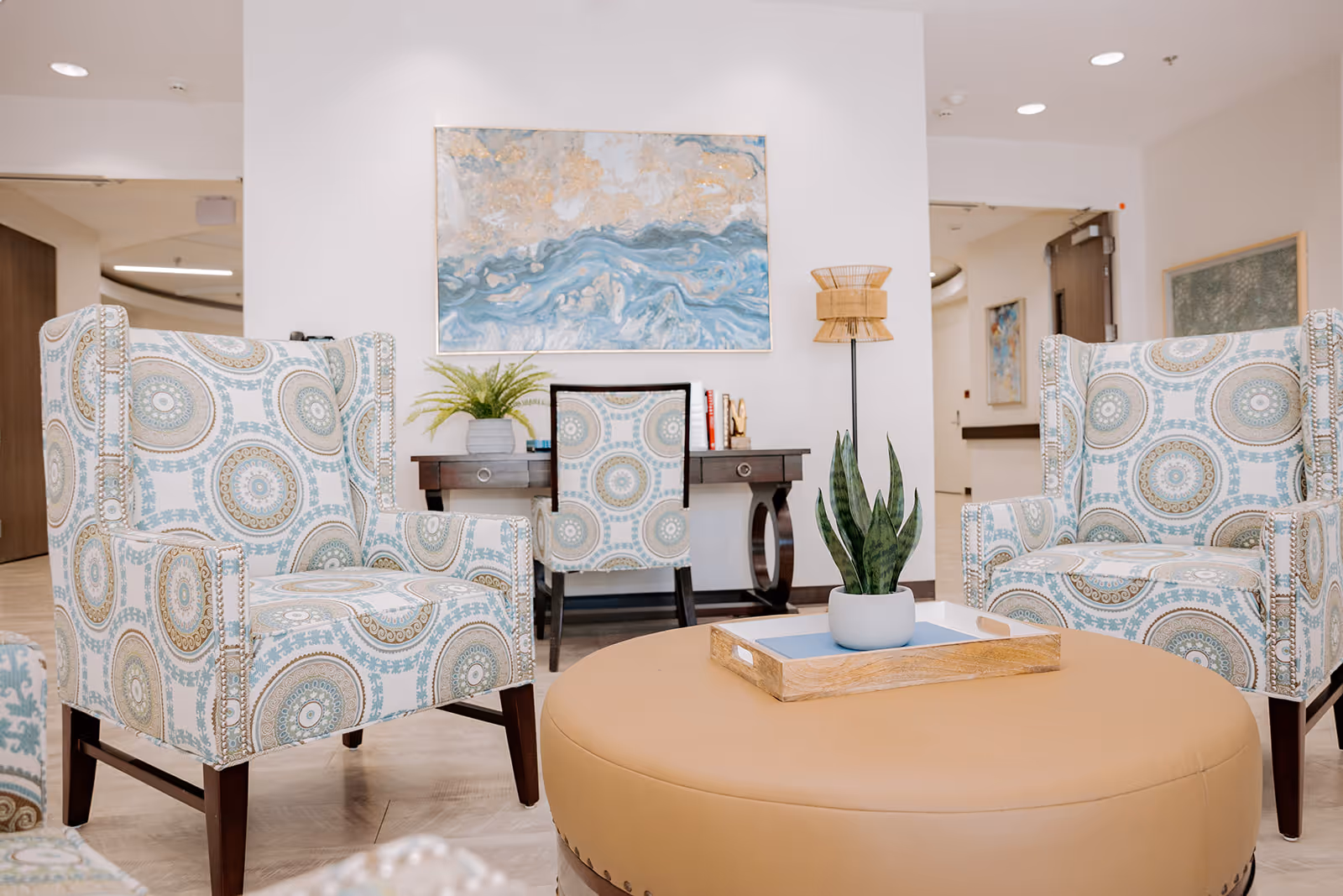 A cozy seating area in a senior living facility featuring two patterned armchairs with circular designs, a round tan ottoman with a wooden tray and a potted plant on top, a small desk with a matching chair, decorative books, a plant, a floor lamp, and abstract artwork on the wall.