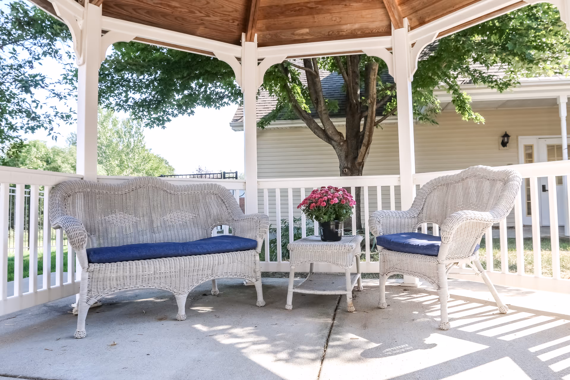 A white wicker loveseat and chair with blue cushions sit under a wooden gazebo. A small wicker table between them holds a pot of pink flowers. The gazebo is surrounded by white railings, with a tree and a beige building visible in the background.