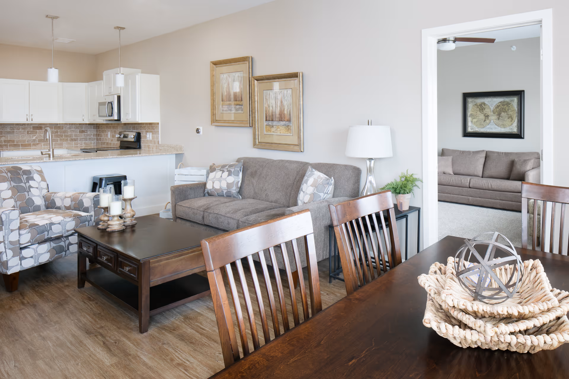 A bright and modern living area with a gray sofa, patterned armchair, and a dark wooden coffee table with candles. The room has light-colored walls with two framed pictures and a side table with a lamp and plant. In the background, there is a kitchen with white cabinets, a microwave, and a stove. A dining table with wooden chairs and decorative baskets is in the foreground. Through an open doorway, another room with a brown sofa and a framed world map is visible.