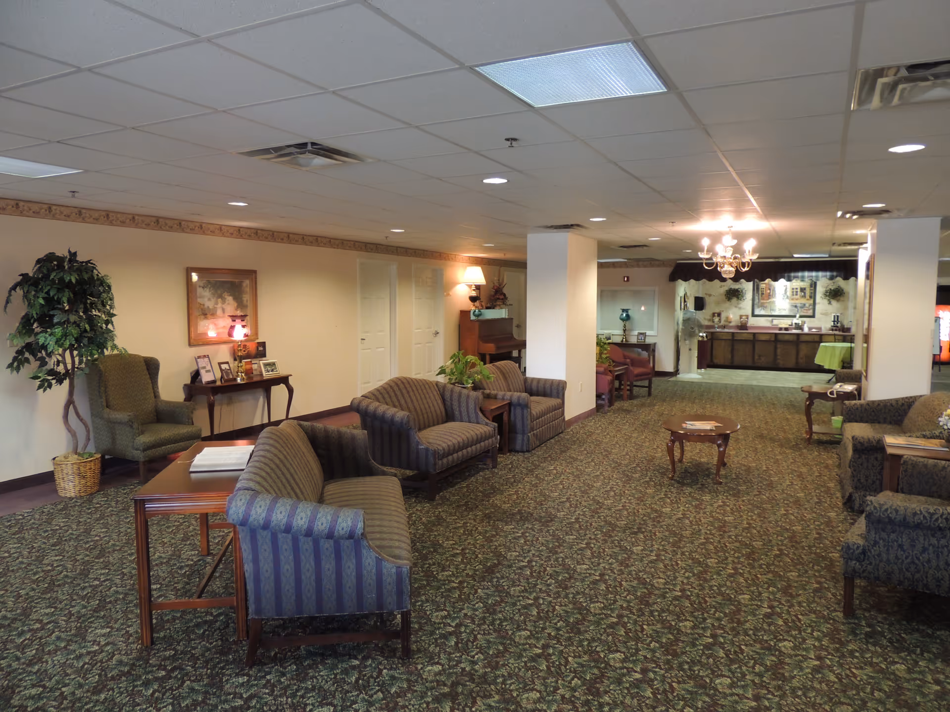 Spacious assisted-living lobby/lounge with upholstered chairs and sofas, tables, plants, and a reception area in the background.