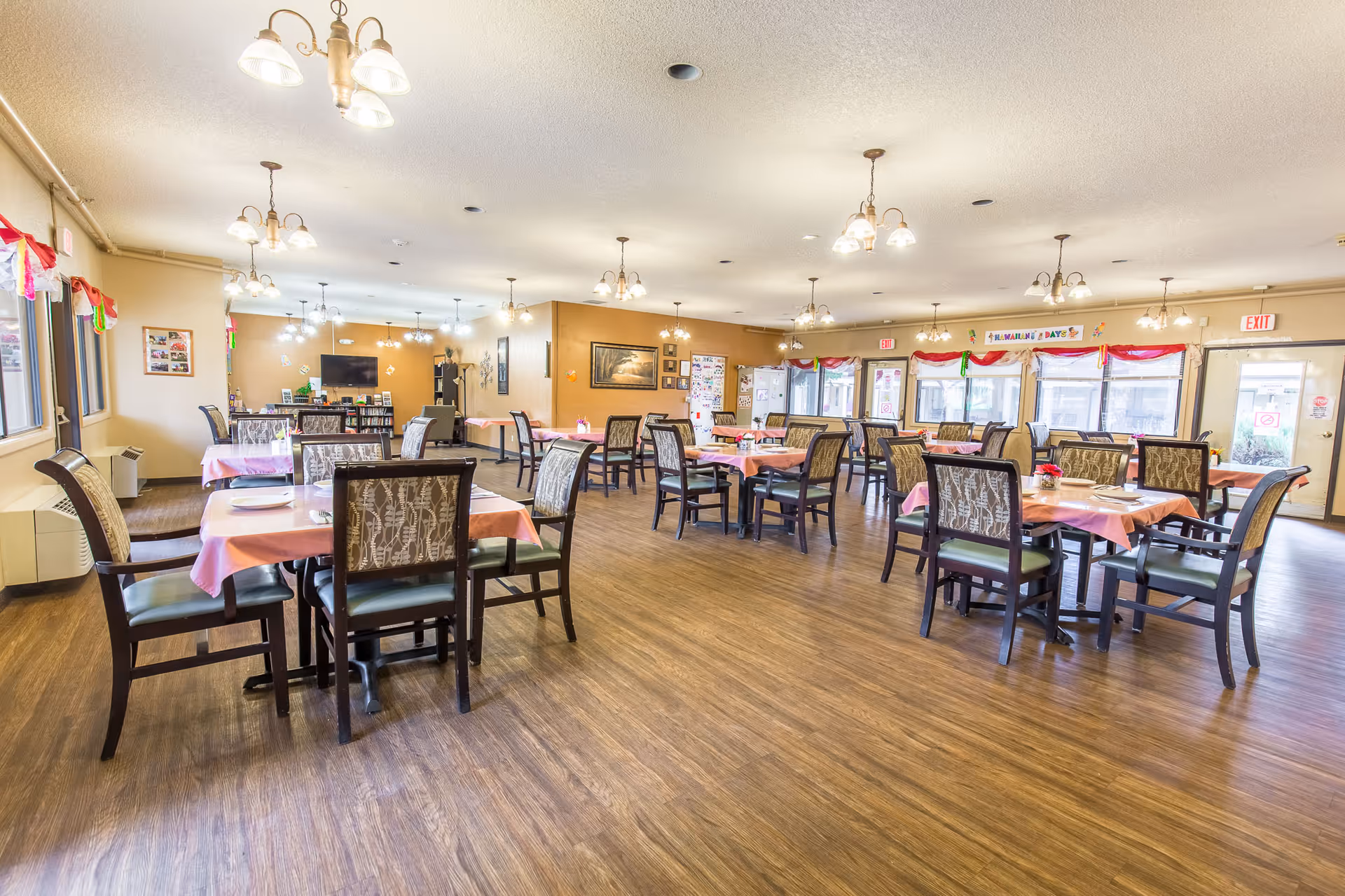 Spacious dining room with multiple tables and chairs set with pink tablecloths, wood flooring, and hanging light fixtures.