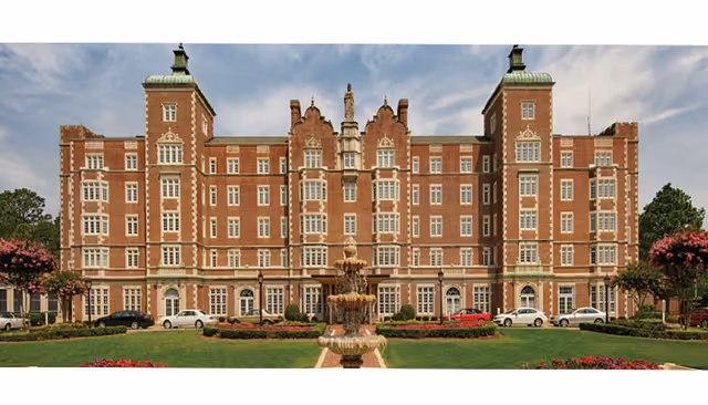 Front exterior view of a large, stately brick building with multiple windows and decorative architectural details. In front of the building is a multi-tiered fountain surrounded by a well-maintained lawn and flower beds. Several cars are parked along the driveway in front of the building.