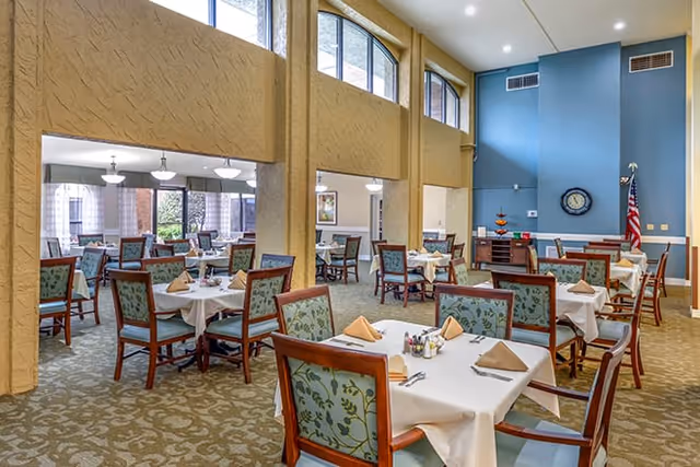 A spacious dining room with multiple tables covered in white tablecloths and set with beige napkins, silverware, and condiments. The room features high ceilings, large windows, and a blue accent wall with a clock and an American flag. The chairs have wooden frames and green patterned upholstery.
