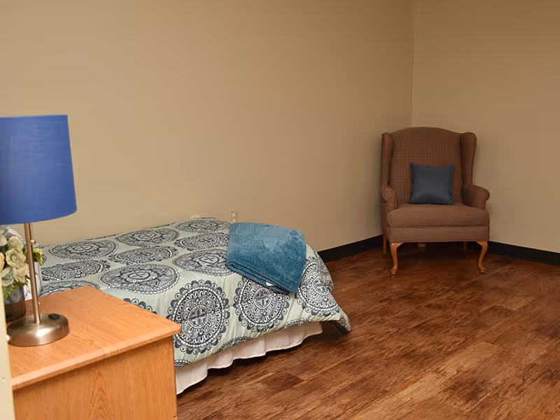 A simple bedroom with a single bed covered in a patterned bedspread and a folded blue blanket on top. There is a wooden nightstand with a blue lamp and a small flower arrangement. In the corner, there is a brown upholstered armchair with a blue cushion. The room has wooden flooring and plain beige walls.