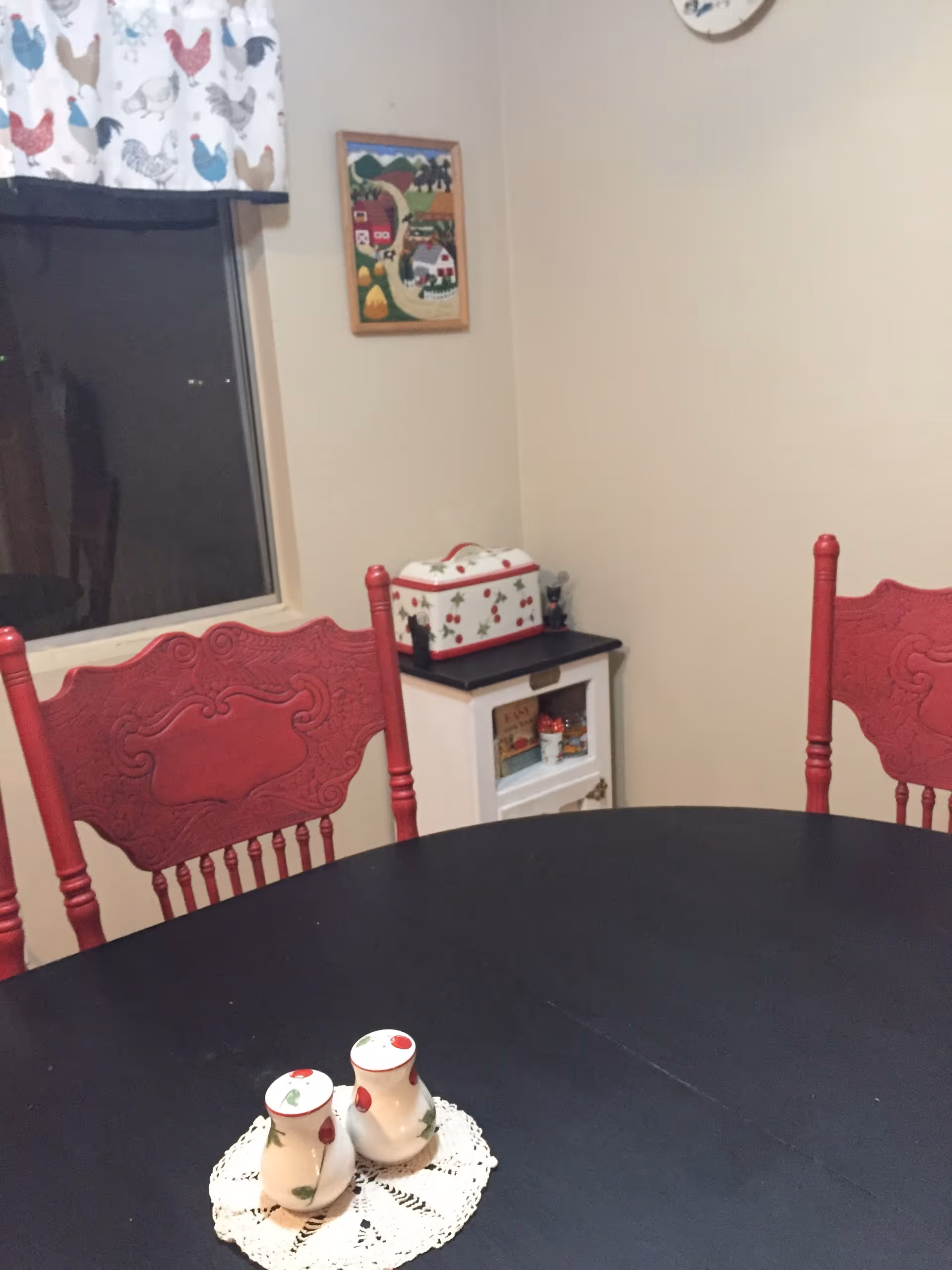 A dining area with a black oval table and two red wooden chairs with ornate backs. On the table are two ceramic salt and pepper shakers on a small white doily. In the background, there is a window with a curtain featuring a chicken pattern, a small white cabinet with a decorative box on top, and a framed picture on the wall.