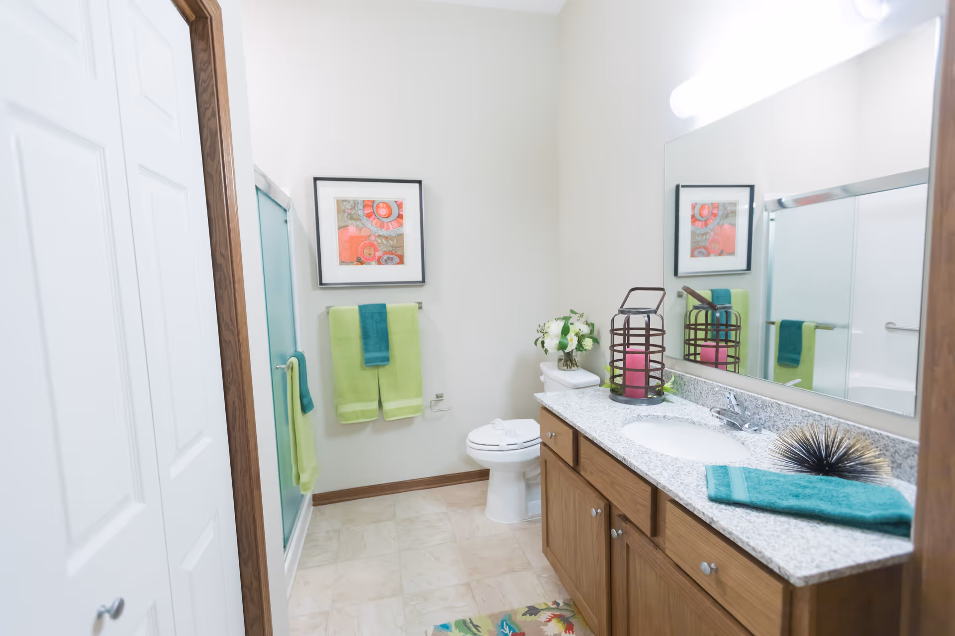 A clean and bright bathroom with a white toilet, a granite countertop with a sink, wooden cabinets, and a large mirror above the sink. There are green and teal towels hanging on the wall and shower door. A framed abstract artwork is hung on the wall above the towels. Decorative items including a candle holder and a small plant are placed on the countertop.