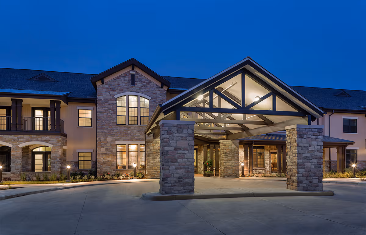 Exterior front view of Spring Creek Village facility at dusk, featuring a large covered entrance with stone pillars and warm interior lighting visible through windows.