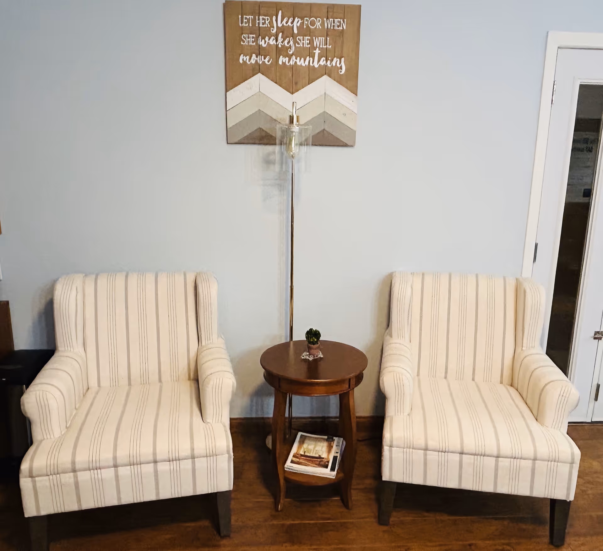 Two beige striped armchairs with wooden legs are placed on either side of a small round wooden table with a small plant on top and magazines on the lower shelf. Behind the table is a tall floor lamp and a wooden wall decoration with the quote 'Let her sleep for when she wakes she will move mountains.' The setting appears to be a cozy indoor seating area with light blue walls and wooden flooring.