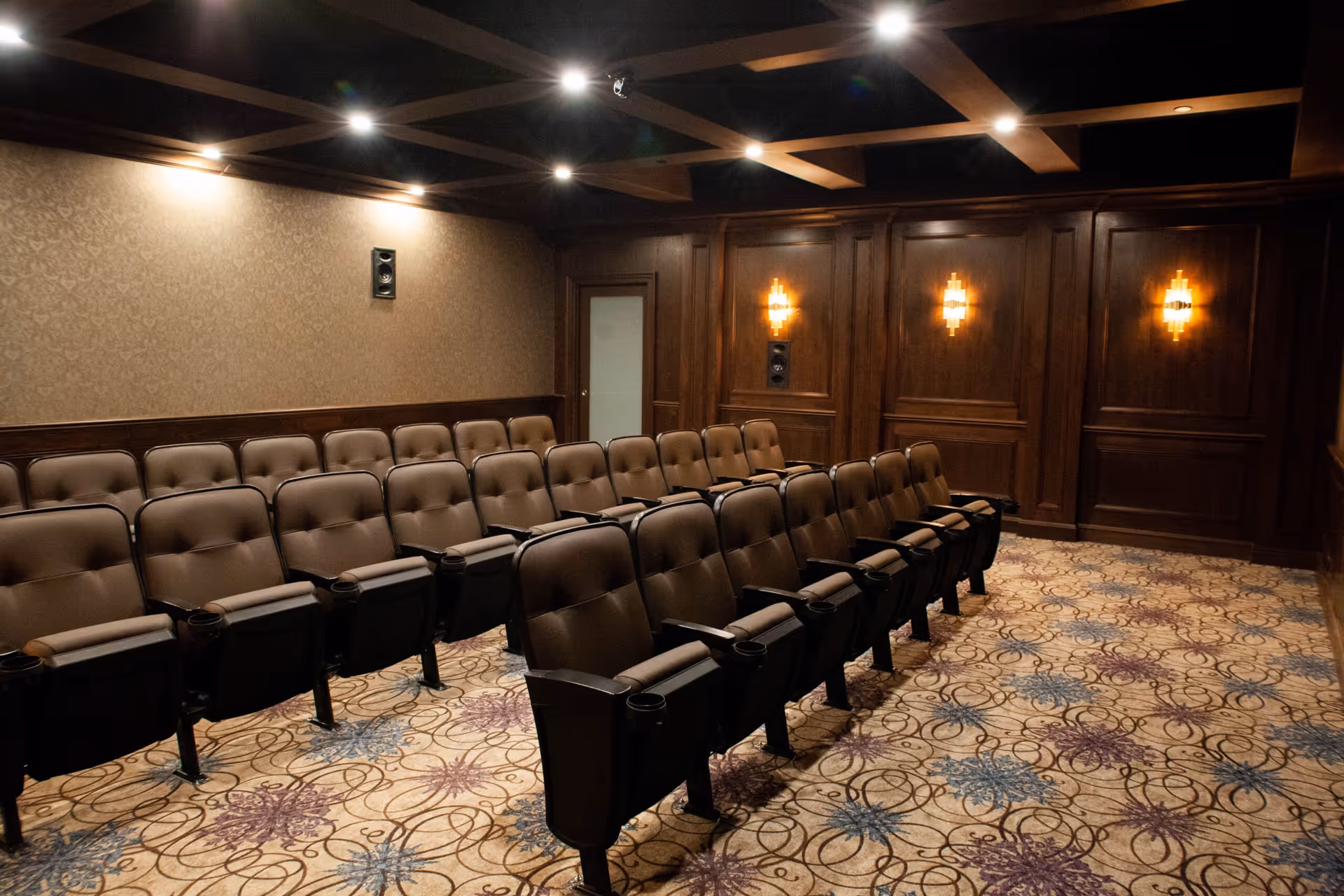 A small wood-paneled theater room with rows of upholstered seats, patterned carpet, and wall sconces.
