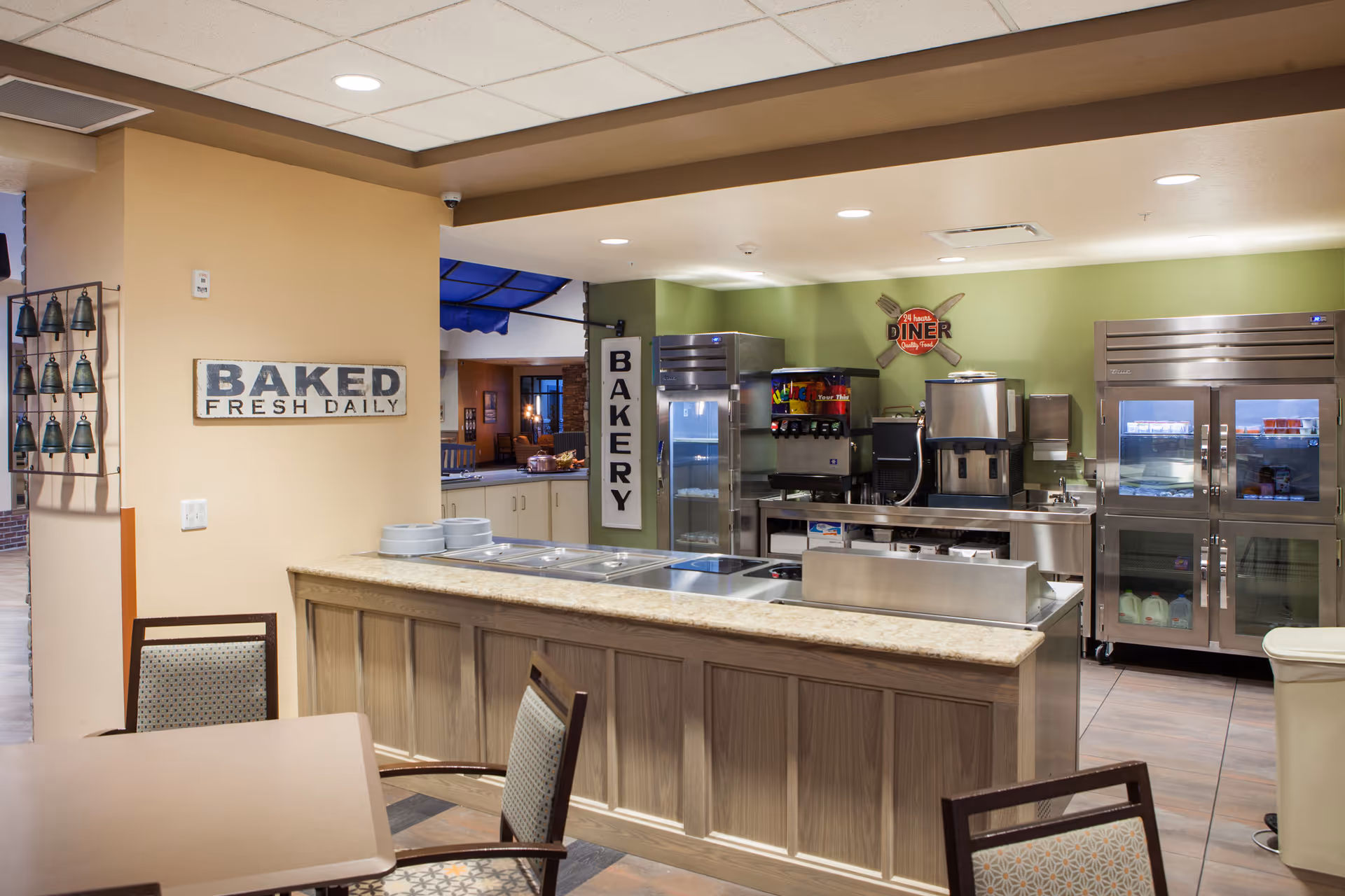 Interior view of a bakery and food service area with a counter, beverage dispensers, refrigerators, and signs that read 'BAKED FRESH DAILY' and 'BAKERY'. There are tables and chairs in the foreground and a green wall with a diner sign in the background.