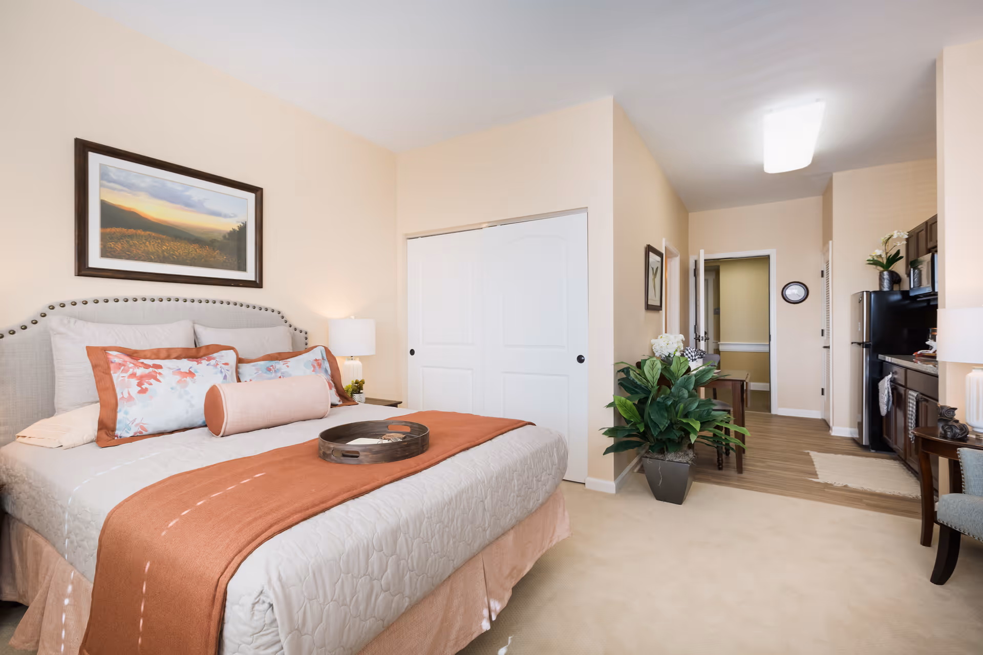 A cozy bedroom in an assisted living facility featuring a neatly made bed with beige and orange bedding, decorative pillows, and a tray on top. The room has beige walls, a framed landscape painting above the bed, a bedside lamp, a large potted plant, and a small kitchen area with dark cabinets and a refrigerator visible in the background.