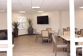 Communal dining/lounge area with multiple tables and chairs, a wall-mounted TV, and potted plants.
