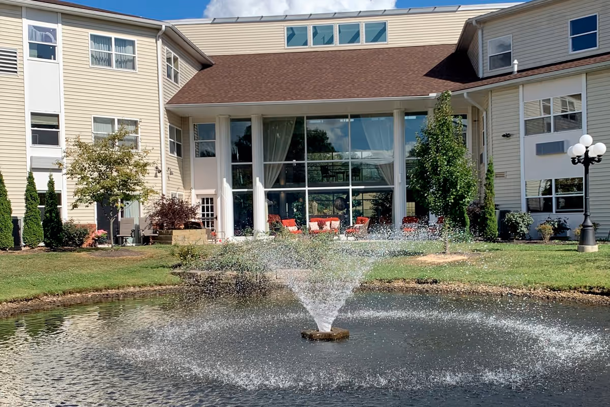 Exterior view of a multi-story retirement facility building with beige siding and large windows. In front of the building is a pond with a water fountain spraying water upwards, surrounded by green grass, trees, and shrubs. There are red chairs visible inside the building through the large windows.