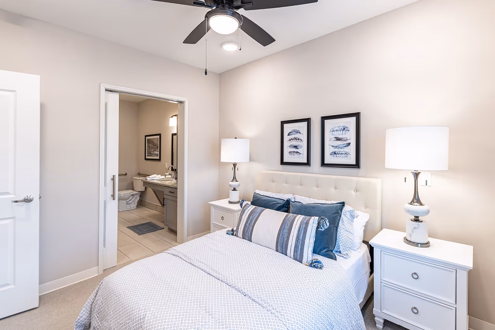 A neatly made bed with white and blue bedding in a senior living bedroom. The bed has a tufted headboard and is flanked by two white nightstands, each with a lamp. Above the bed are two framed pictures of feathers. A ceiling fan with a light is visible on the ceiling. An open door reveals an adjacent bathroom with a toilet, sink, and mirror.