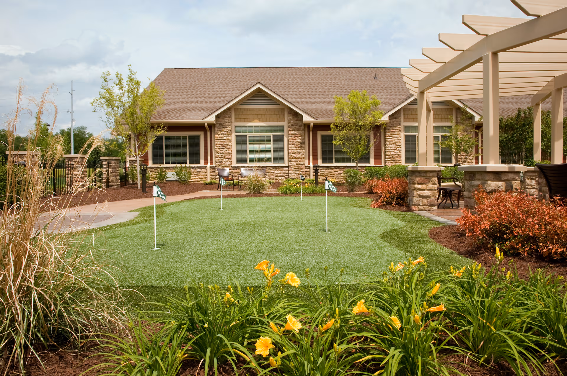 Outdoor garden area with a putting green featuring four small flags, surrounded by yellow flowers and various shrubs. In the background, there is a single-story building with stone and siding exterior, multiple windows, and a pergola-covered patio area with seating.