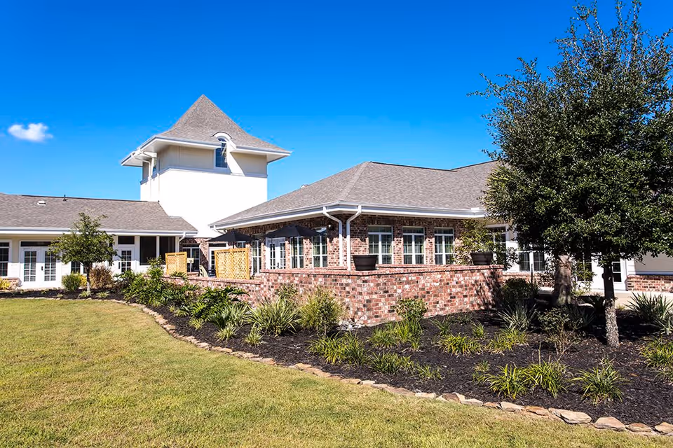 Exterior view of The Verandah Retirement Community building with a brick facade, multiple windows, a tower-like structure, and a landscaped garden with trees and shrubs under a clear blue sky.