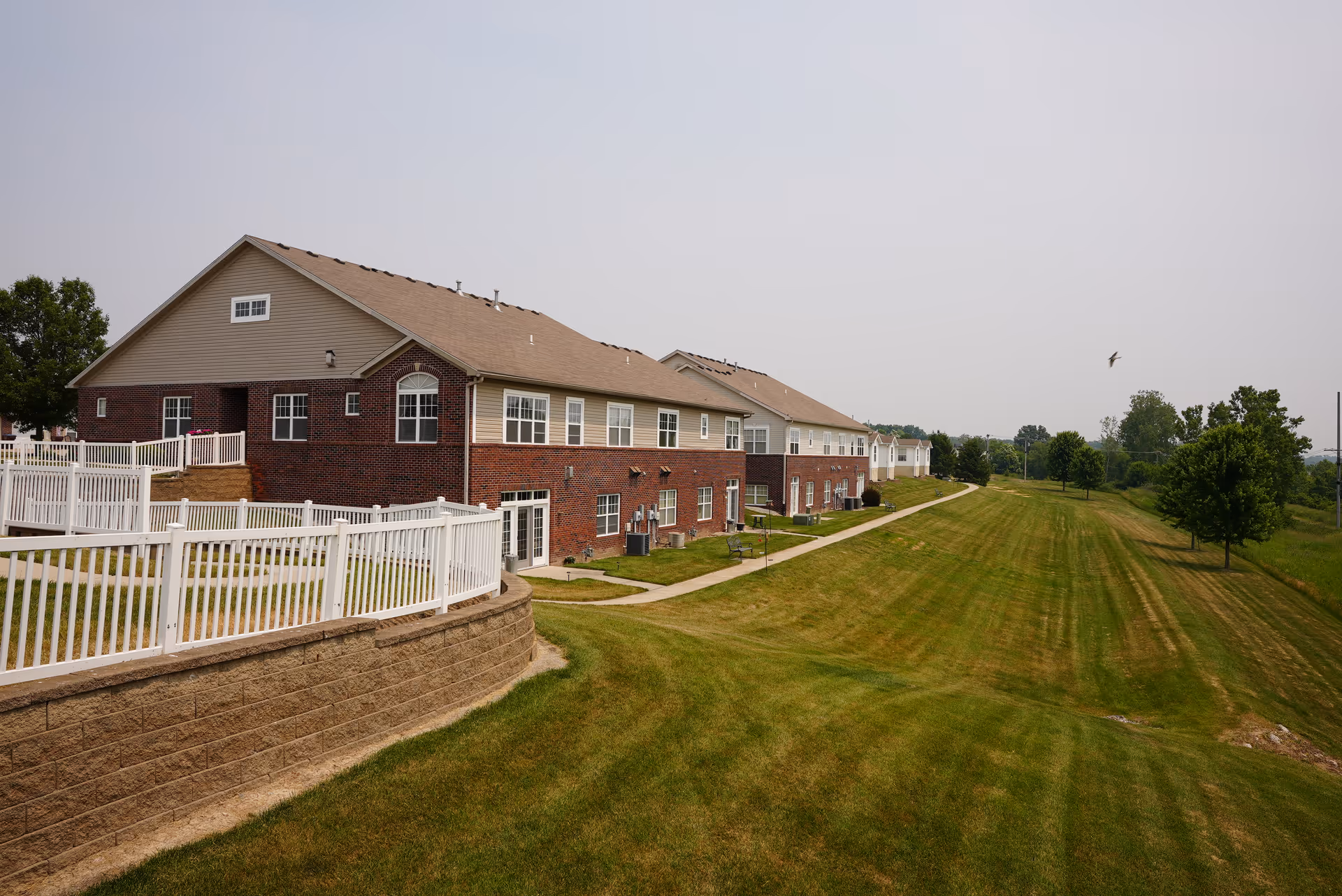 Long brick assisted-living building with white fencing, a paved walkway and a large mown lawn under a pale sky.