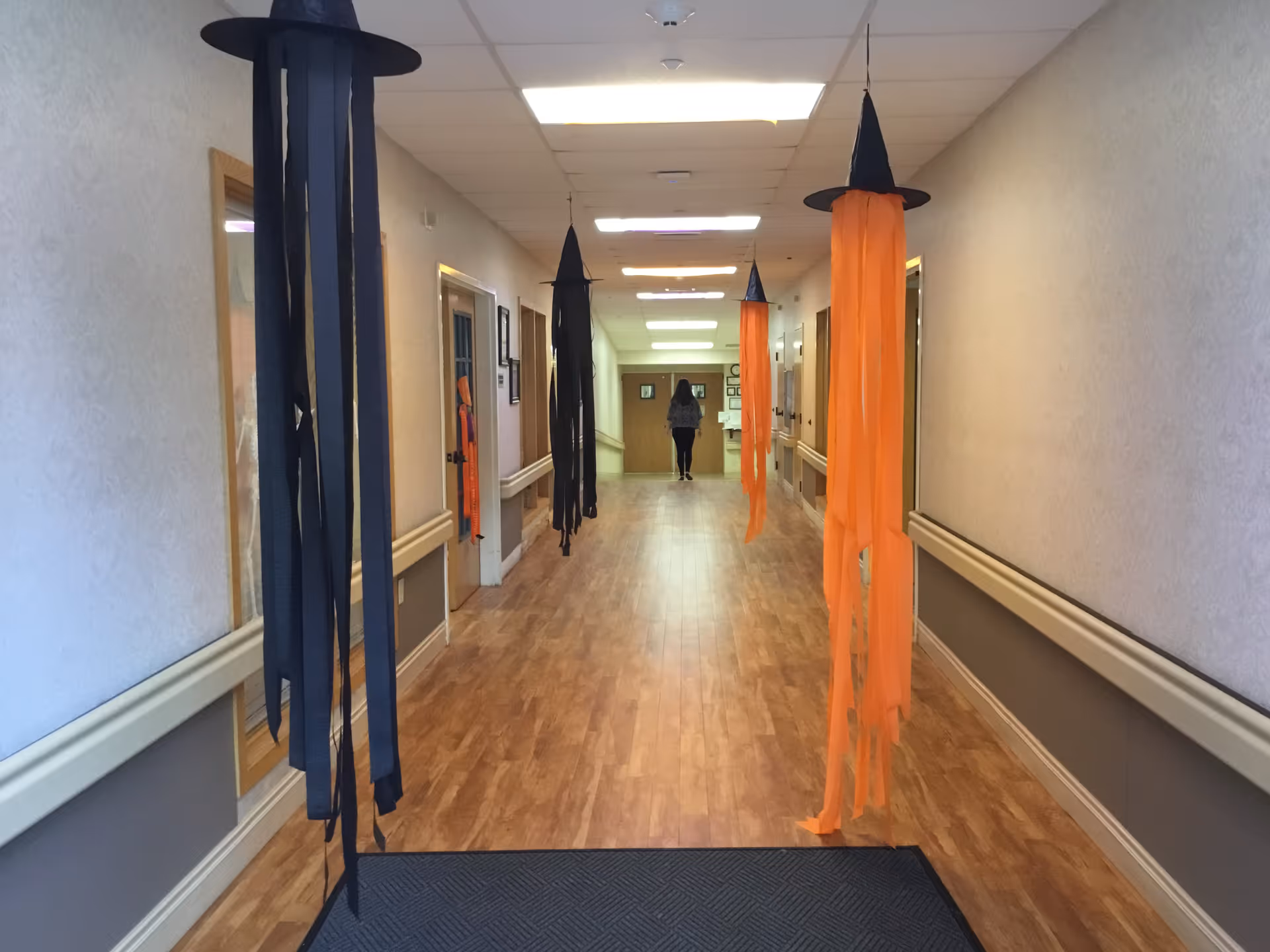 A long interior hallway in a care facility decorated with hanging black and orange witch-hat streamers and a person walking away at the far end.