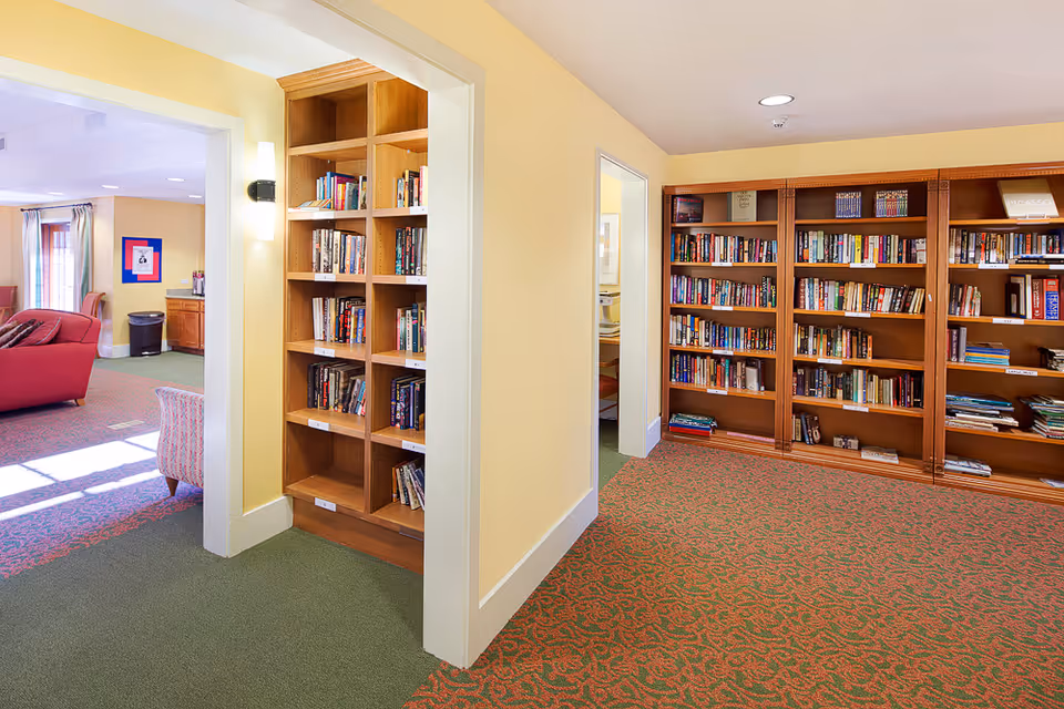 Interior view of a senior living facility showing a cozy reading area with wooden bookshelves filled with books. The room has yellow walls and a patterned carpet. To the left, there is an open doorway leading to a living room area with red and patterned armchairs and natural light coming through windows.