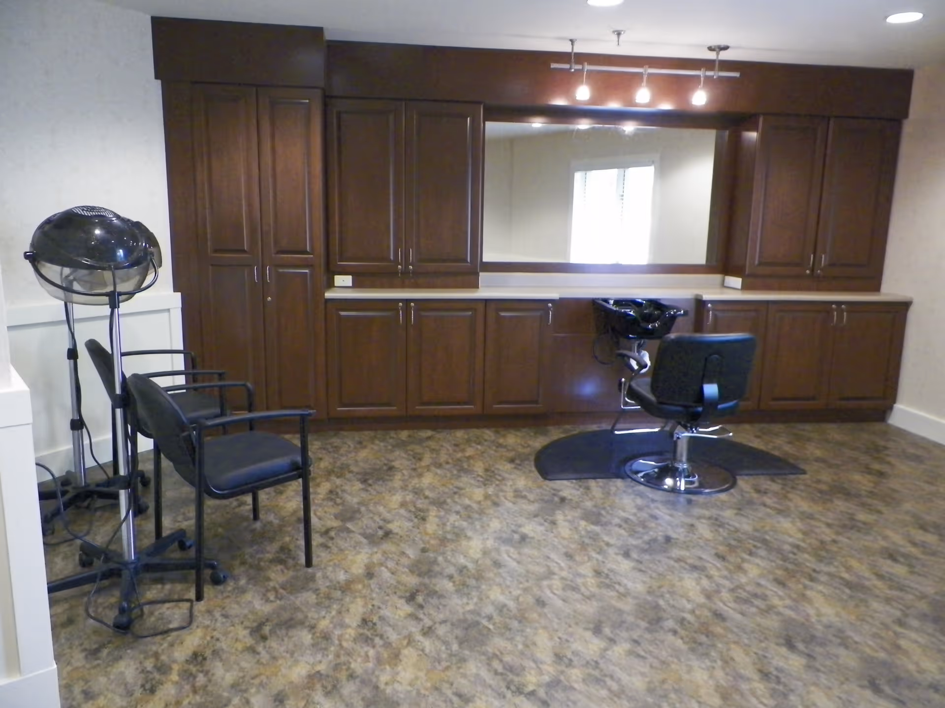Interior view of a hair salon area in a senior living facility featuring a black salon chair in front of a large mirror with wooden cabinets above and below the countertop. To the left, there is a black hair dryer on a stand and two black chairs. The floor has a marbled pattern.