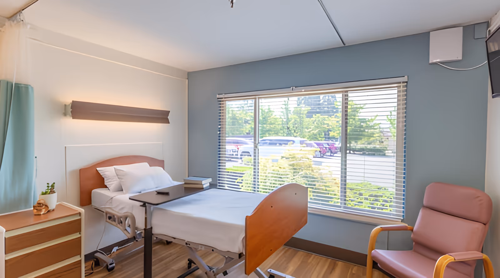 A bright nursing facility bedroom with a hospital bed, a small overbed table with books and a remote, a wooden dresser with a small plant, a large window with blinds showing a parking lot outside, and a comfortable pink armchair.