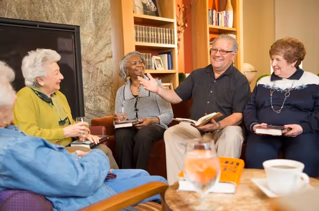 A group of five elderly people sitting in a cozy living room, engaged in a lively conversation. They are seated on comfortable chairs and a sofa around a coffee table with drinks and books. Bookshelves and a fireplace are visible in the background.