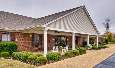 Exterior view of a single-story brick building with a covered porch supported by white columns. The porch has white chairs and potted plants, with neatly trimmed bushes and a paved walkway in front. The sky is overcast.