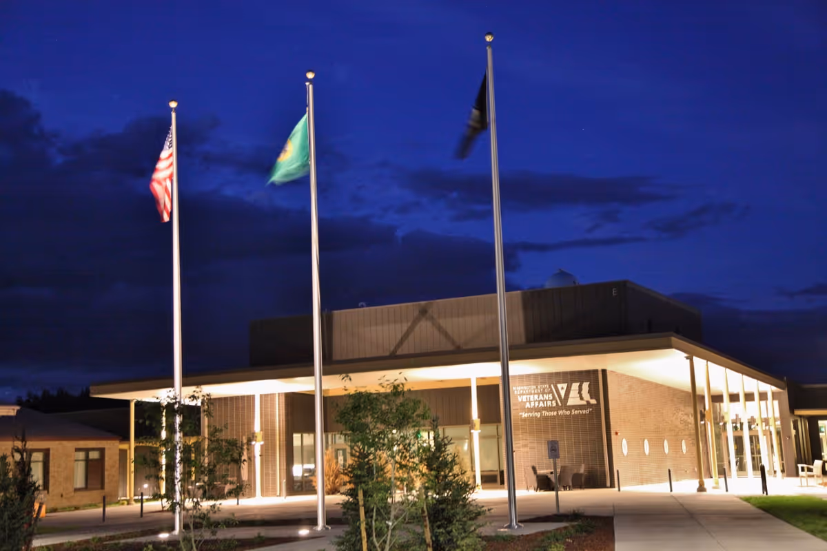 Exterior view of the Walla Walla Veterans Home building at night with three flagpoles displaying flags in front. The building is illuminated with lights and has a modern design with a covered entrance.