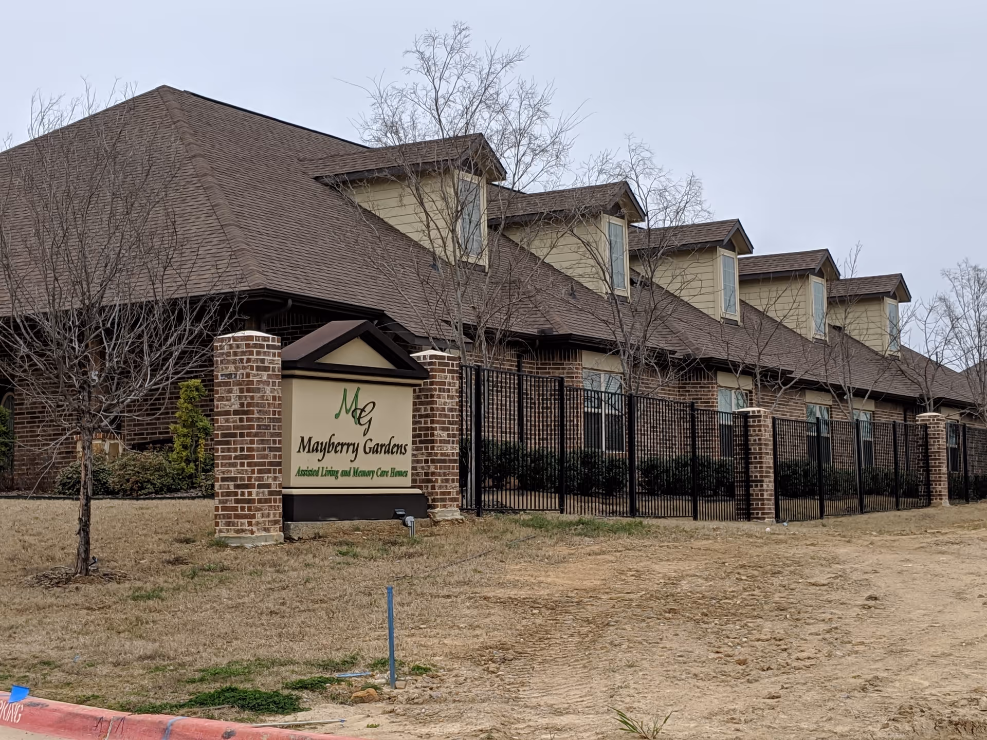 Exterior view of Mayberry Gardens Assisted Living and Memory Care Homes building with a brick facade, multiple dormer windows, a fenced area, and a sign displaying the facility's name in front of the building.