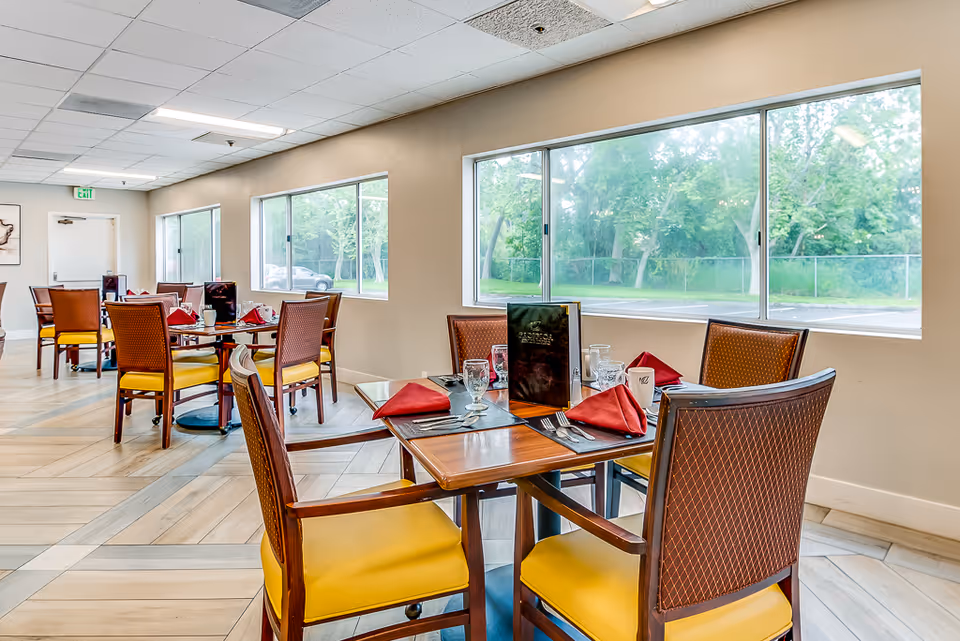 A bright dining room with multiple wooden tables and chairs featuring yellow cushions. Each table is set with red folded napkins, glassware, and menus. Large windows on the right side provide a view of green trees outside.