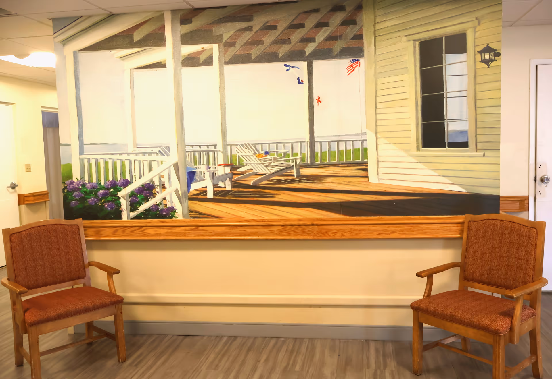 Two wooden armchairs sit in front of a large mural depicting a sunlit porch on an interior wall.