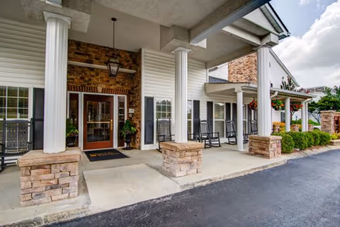 Entrance of a senior living facility with a covered porch supported by white columns and stone bases. There are rocking chairs lined up along the porch and hanging flower baskets. The building exterior features brick and white siding with large windows and a double glass door entrance.