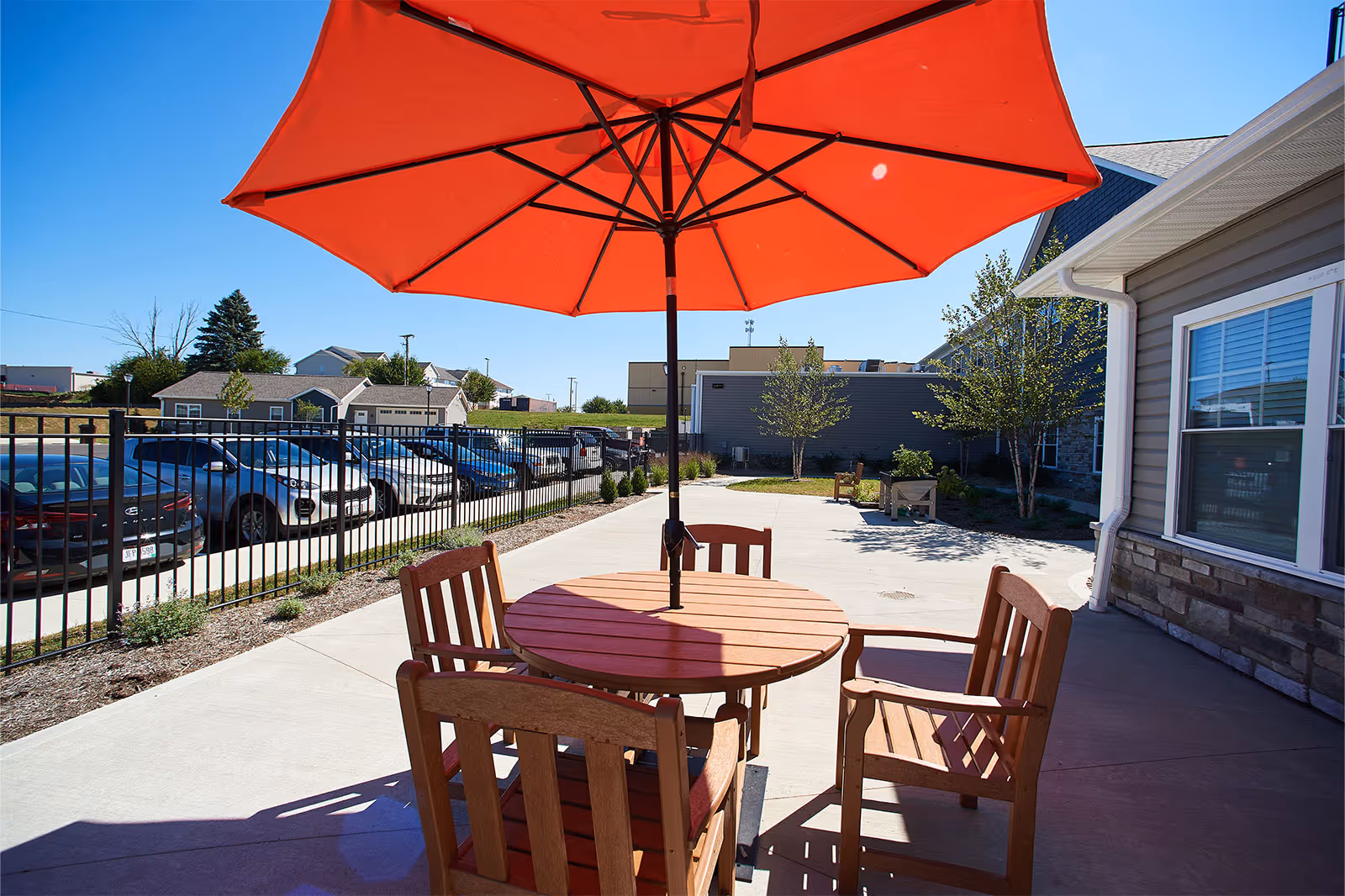 Outdoor patio area with a round wooden table and four wooden chairs under a large red umbrella. The patio is adjacent to a building with beige siding and stone accents. There is a black metal fence separating the patio from a parking lot with several parked cars. Trees and shrubs are planted along the patio and building.