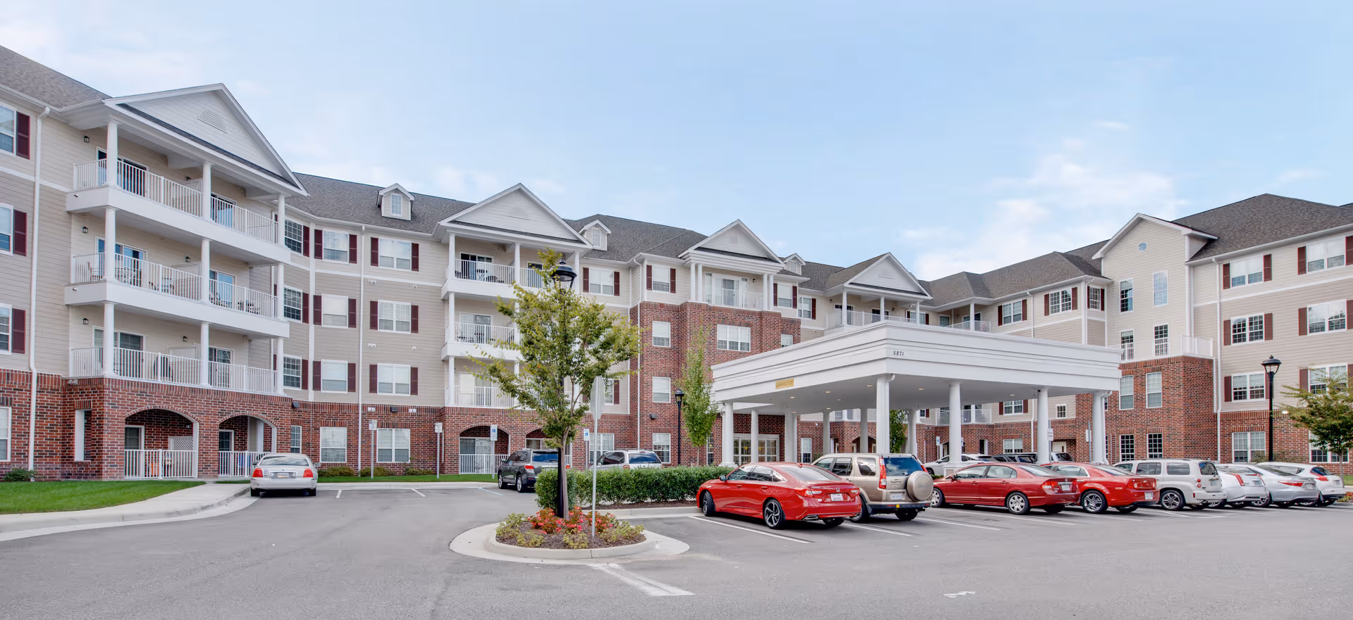 Front exterior of a multi-story senior living building with a covered entrance, balconies, and parked cars in the driveway.
