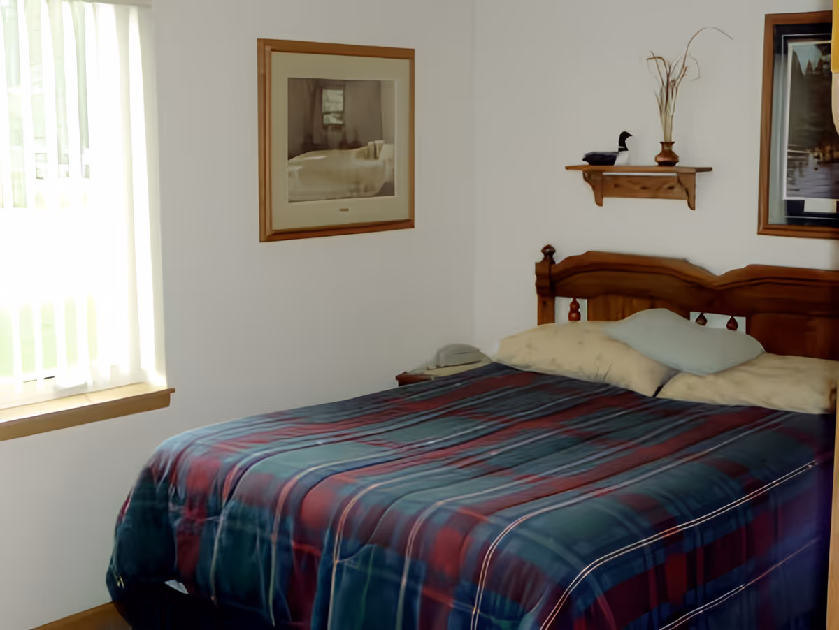 A simple bedroom with a wooden headboard, a plaid bedspread and pillows, wall art, a small shelf with decor, and a window with vertical blinds.