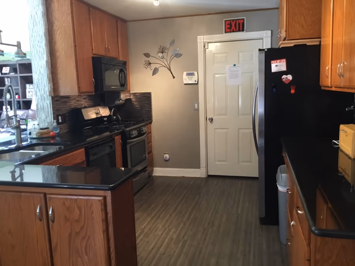A kitchen with wooden cabinets, black countertops, a stainless steel stove with a black microwave above it, a black refrigerator with magnets and a 'File of Life' sticker, a sink with a modern faucet, and a door with an exit sign above it. The floor is wood-patterned, and there is a decorative metal wall art piece on the wall near the door.