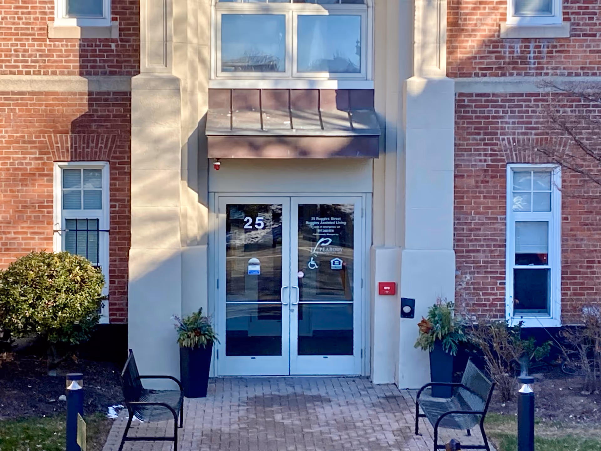 Entrance to a brick building with double glass doors numbered 25. There are two black benches and two tall planters with greenery on either side of the entrance. The building has large windows and a small awning above the doors.