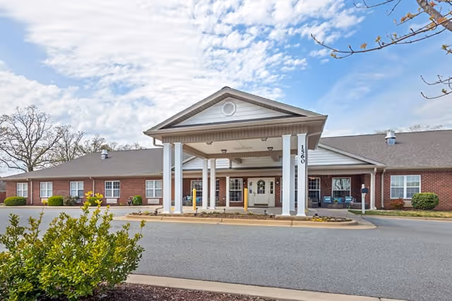 Front exterior view of a single-story brick building with a covered entrance supported by white columns, a driveway in front, and a partly cloudy sky above.