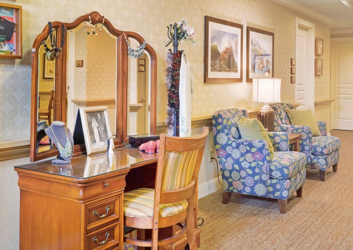 A cozy interior space featuring a wooden vanity table with a tri-fold mirror, adorned with necklaces and a framed black-and-white photo. A wooden chair with striped upholstery is placed in front of the vanity. To the right, there are two floral-patterned armchairs with green cushions, a side table with a lamp, and framed artwork on the wall. The room has beige wallpaper and carpeted flooring.