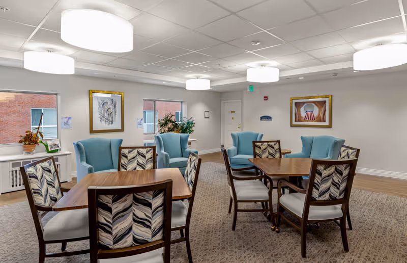 A well-lit common area in a senior living facility with two wooden tables surrounded by chairs featuring patterned upholstery. There are four light blue armchairs arranged near the walls, two windows showing a brick exterior, framed artwork on the walls, and ceiling lights providing illumination.
