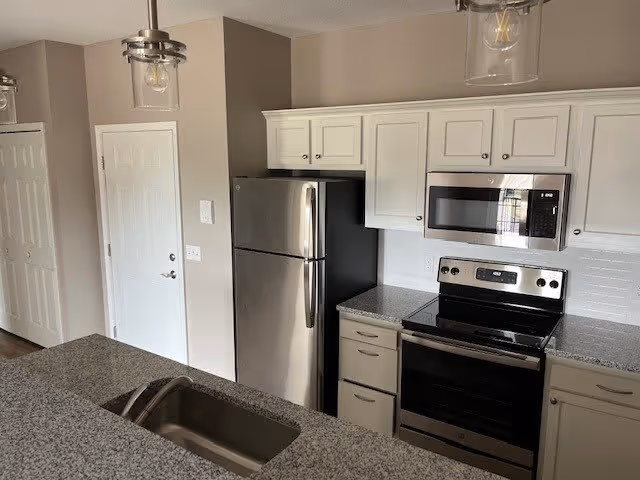 Modern kitchen with stainless steel refrigerator, electric stove with oven, microwave, white cabinets, granite countertops, and a sink in the foreground. The walls are painted beige and there are two pendant lights hanging from the ceiling.