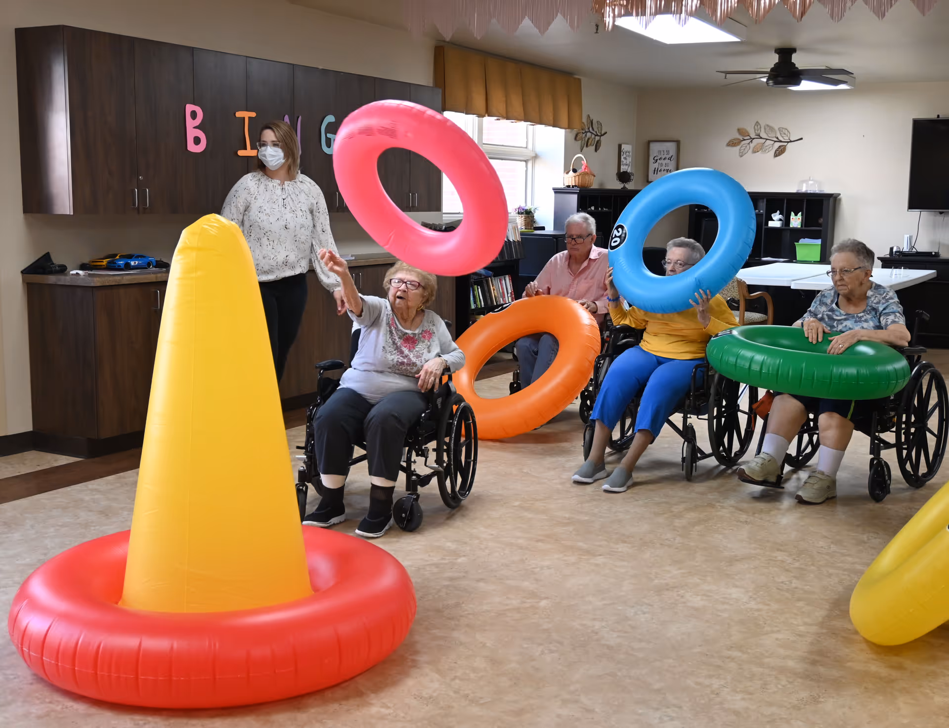 Four elderly women in wheelchairs playing a ring toss game indoors with colorful inflatable rings and a large yellow cone. A caregiver wearing a mask stands nearby watching. The room has wooden cabinets, a ceiling fan, and a TV mounted on the wall.
