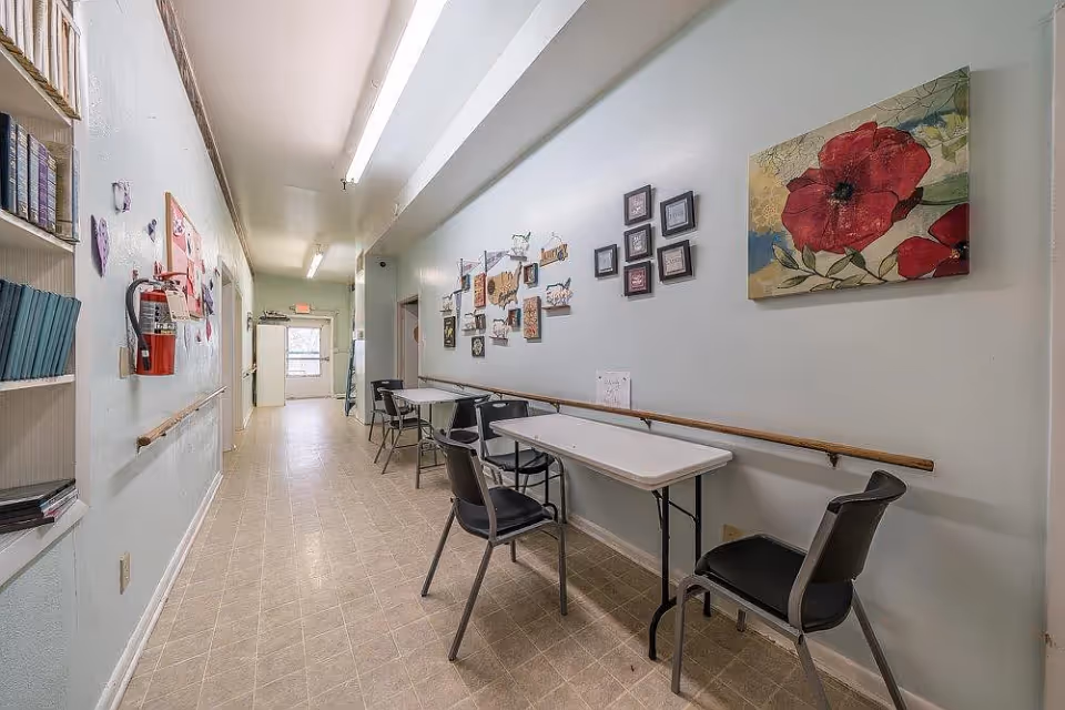 A long hallway in a senior living facility with light-colored walls and tiled floor. On the right side, there are folding tables with black chairs placed against the wall, decorated with various framed pictures and artwork including a large red flower painting. On the left side, there are shelves with books and a fire extinguisher mounted on the wall. The hallway is well-lit with fluorescent ceiling lights and has handrails on both sides.