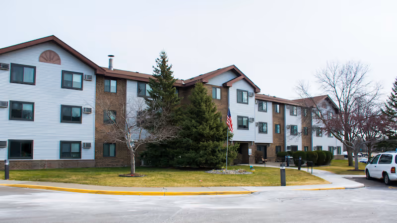 Exterior view of a three-story senior living facility building with white siding and brown brick accents. There are several windows, a few trees including evergreen and leafless deciduous trees, a small lawn area, a flagpole with an American flag, and a parking area with a white vehicle visible on the right side.