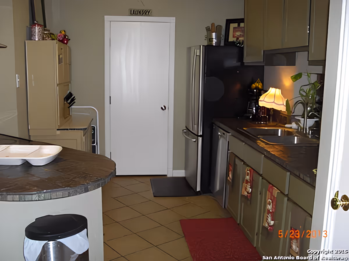Small kitchen interior with an island, stainless steel refrigerator, sink and a lamp on the countertop.
