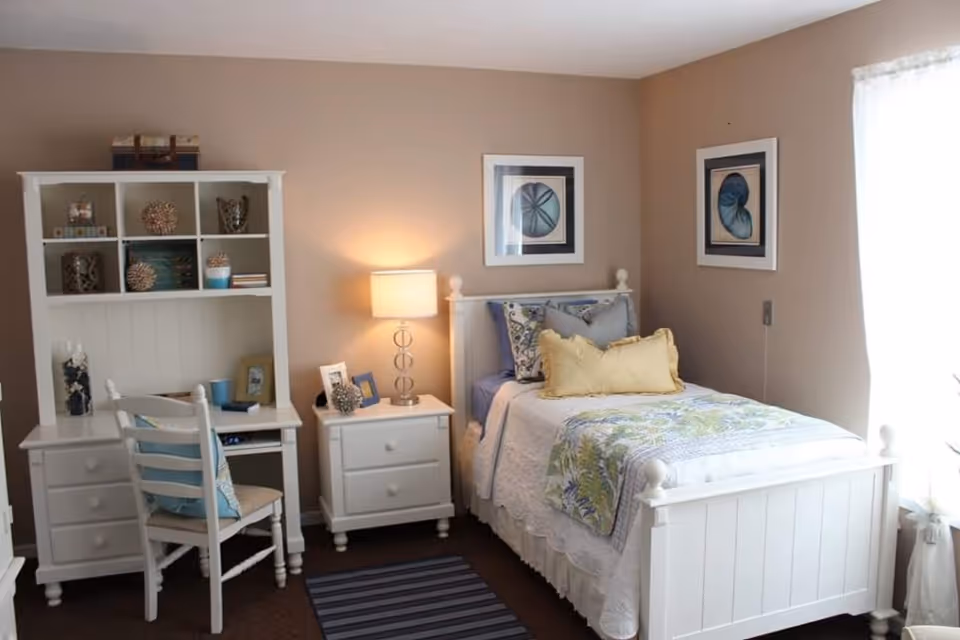 A cozy bedroom with a white wooden bed featuring a floral quilt and multiple pillows. Next to the bed is a white nightstand with a lamp and decorative items. To the left, there is a white desk with shelves holding various decorative objects and a white chair with a cushion. The walls are painted beige, and two framed pictures hang above the bed. A window with sheer curtains allows natural light to brighten the room.
